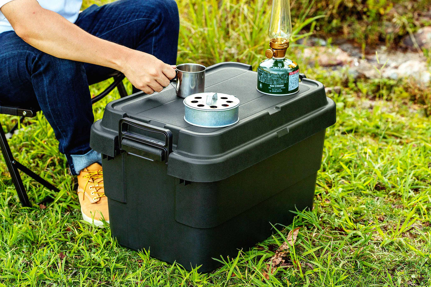 A man sitting on the grass is using a black camping storage box as a table for his camping gears