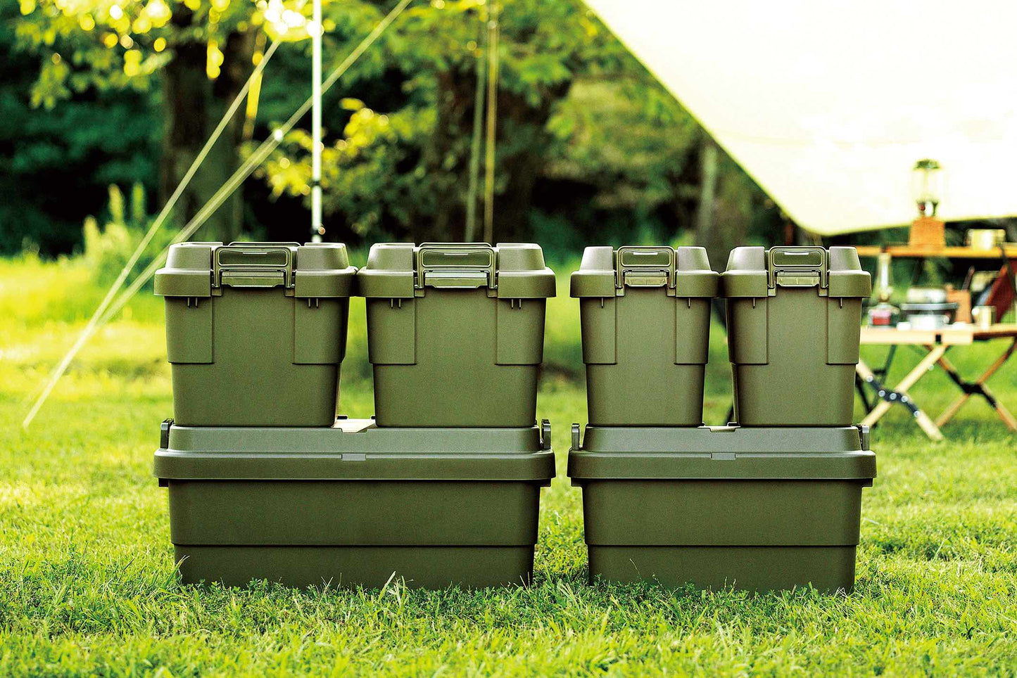 Six green camping storage boxes with different capacities stacking up with their combination and sitting on the grass next to a tent